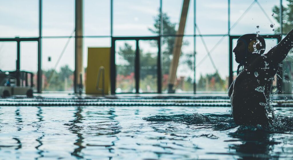 Silhouette of a water polo player mid-action with water splashes inside an indoor pool.
