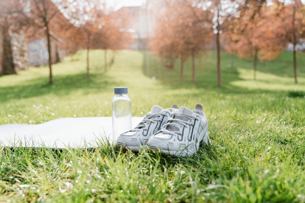 Running shoes, water bottle, and yoga mat on grass in a sunny park setting.